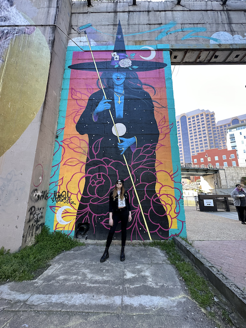 A woman dressed in black standing in front of a giant painting of a witch on a wall